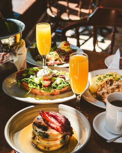 Table set for brunch with avocado on toast, pancakes, and two glasses of mimosas