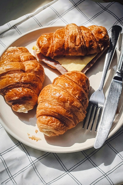 three croissants on a plate, one is filled with ham and cheese, there is a knife and fork beside it