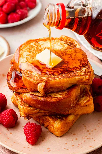 French toast on a plate served with raspberries, butter and maple syrup being poured on it